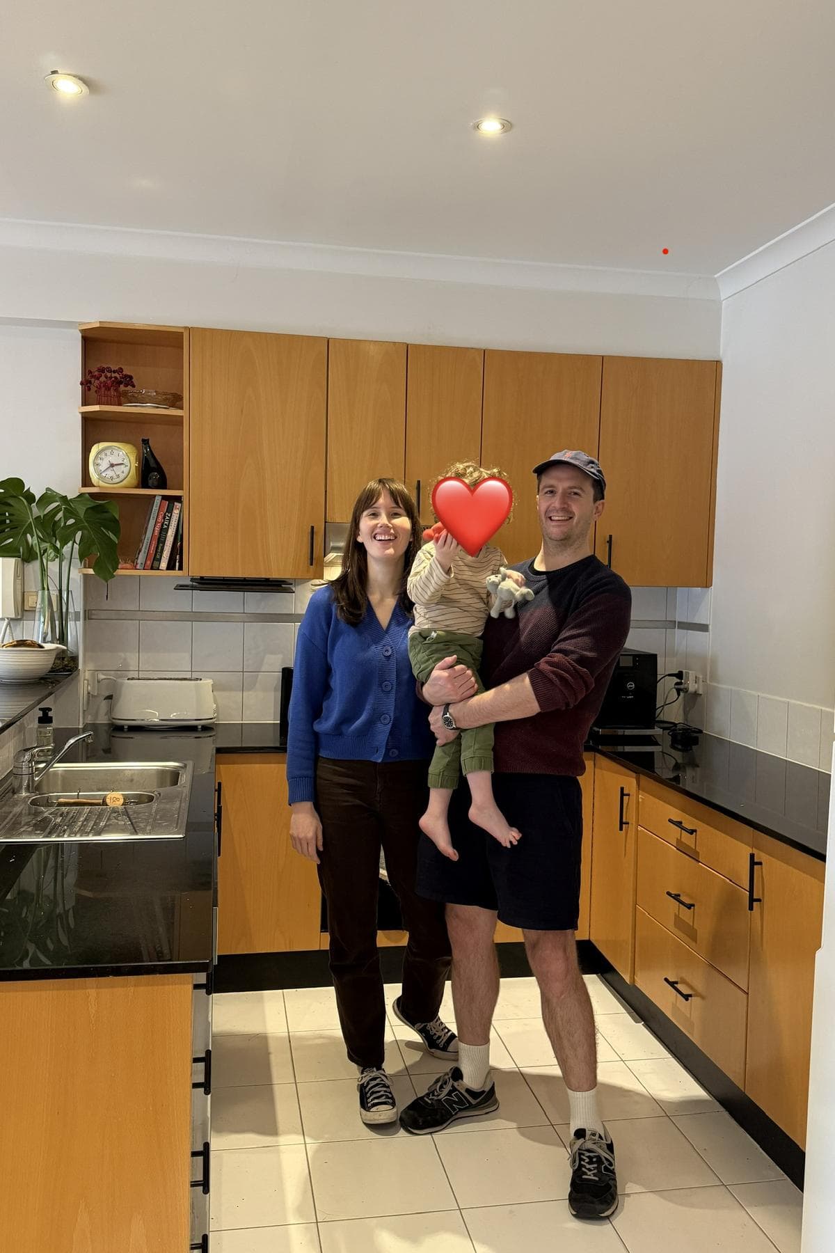 Two adults and a toddler standing in a kitchen with timber cabinets, black benchtops and white tiled floor.