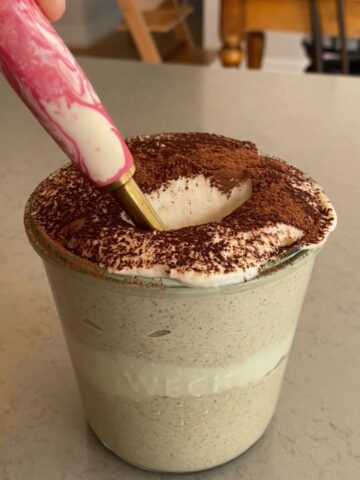 Spoon dipping into creamy tiramisu blended oats topped with whipped cream and a dusting of cocoa powder.