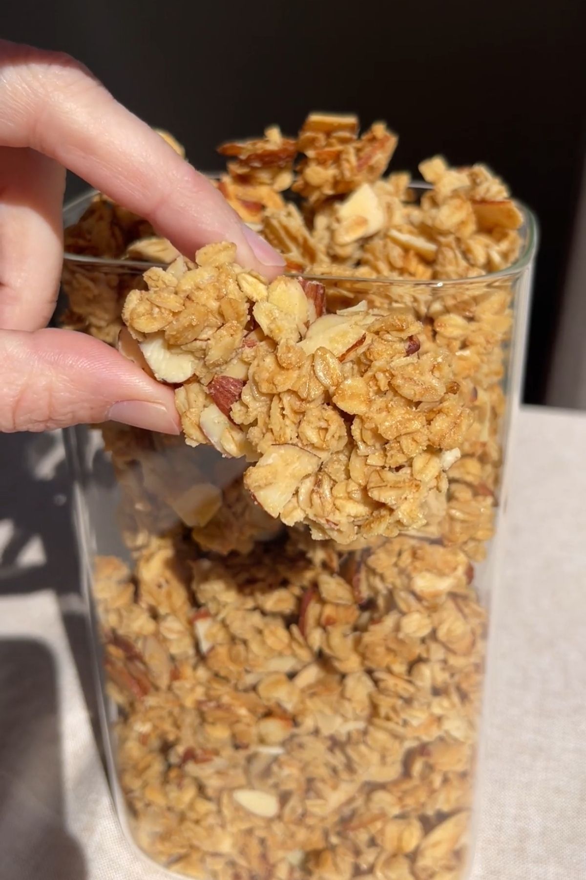 Hand holding a crunchy vanilla almond granola cluster over a container of granola.