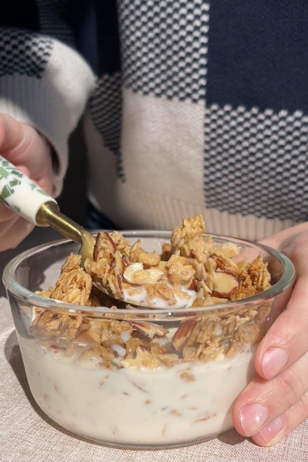 Hands holding a bowl of vanilla almond granola with plant-based milk and a spoon.