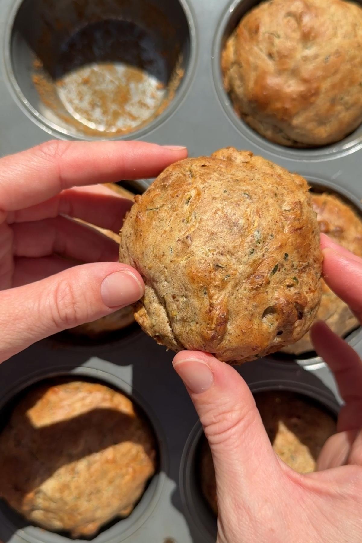 Hands holding a golden brown vegan zucchini muffin over a muffin pan.
