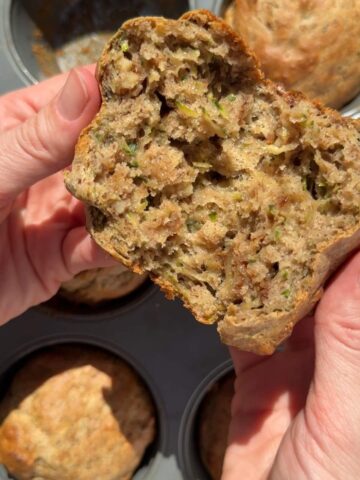 Hands holding a fluffy vegan zucchini muffin with a moist crumb and shredded zucchini visible.