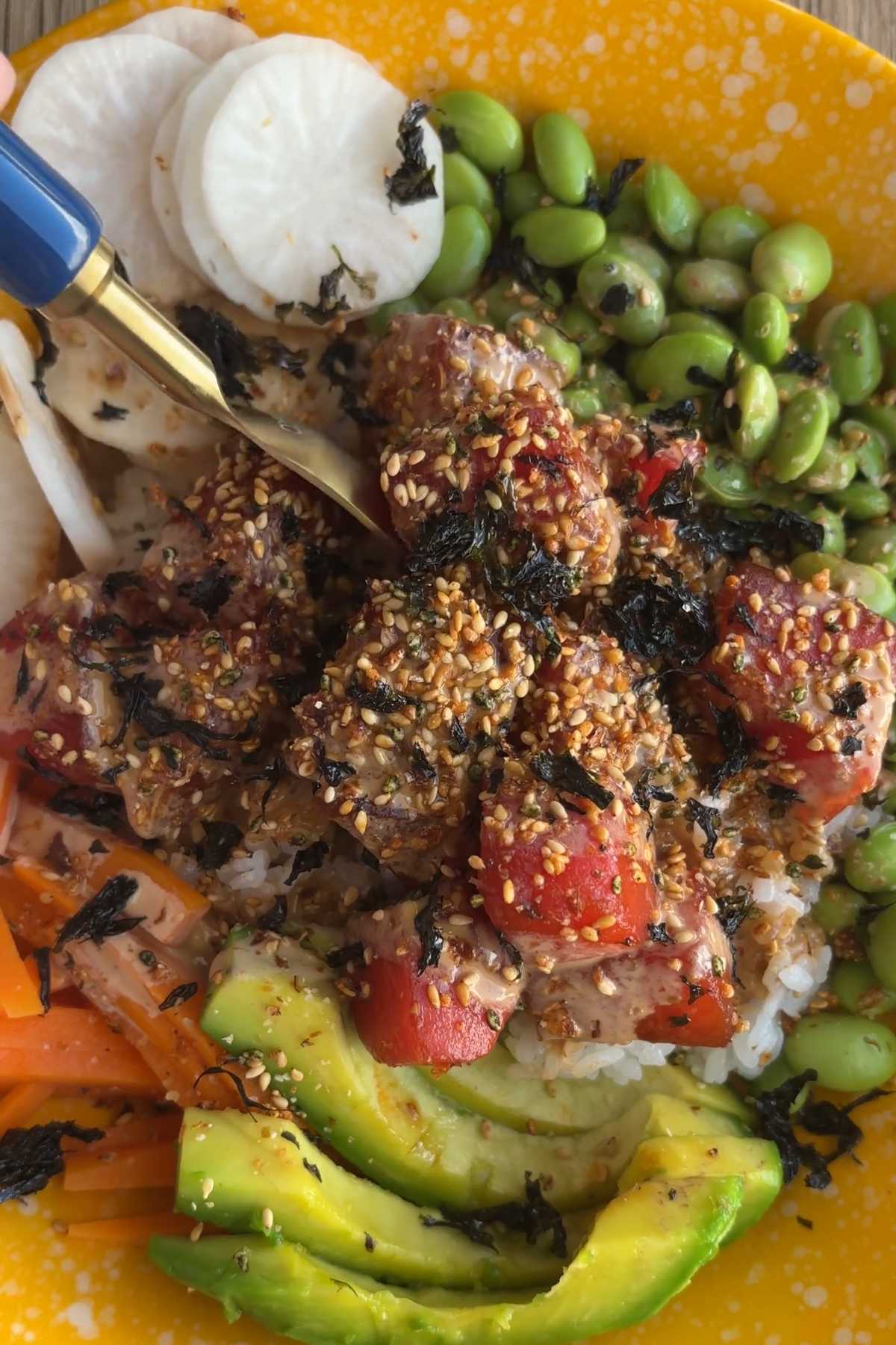 Close-up of a spoon lifting watermelon from a poke bowl topped with sesame seeds and seaweed.
