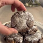 Hand lifting a powdered sugar-dusted chocolate crinkle cookie from a stacked plate.