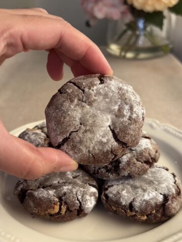 Hand lifting a powdered sugar-dusted chocolate crinkle cookie from a stacked plate.