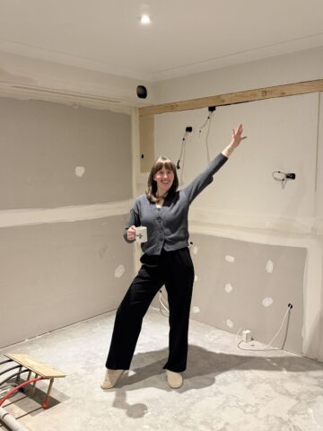 Woman holding a mug and raising one arm in an empty room stripped back for renovation.