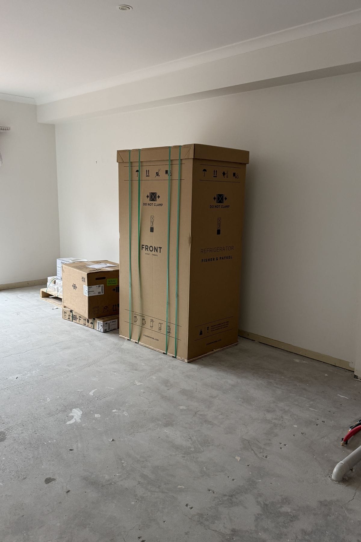 Large boxed refrigerator standing in an empty room beside boxed appliances on a pallet.