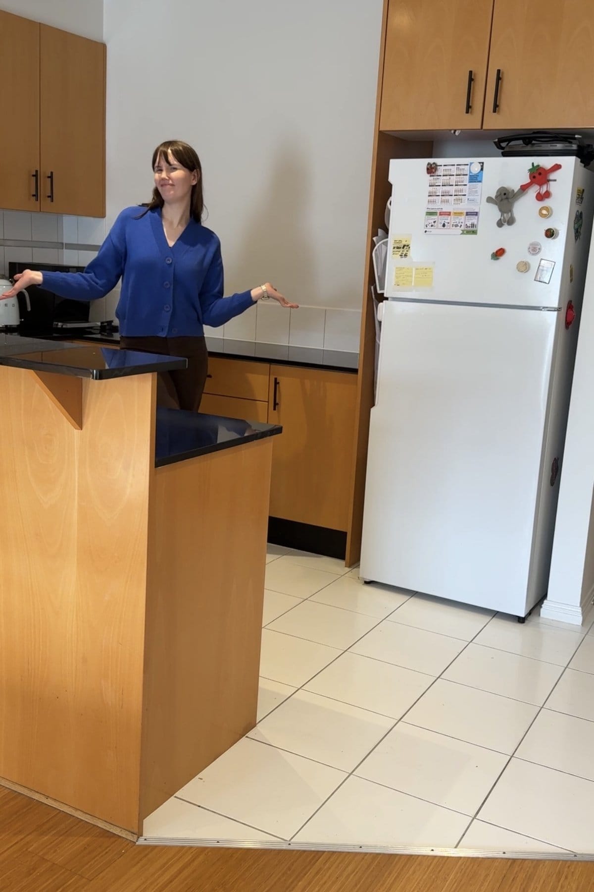 Woman standing in the old kitchen with maple cabinets, black benchtops and white floor tiles.