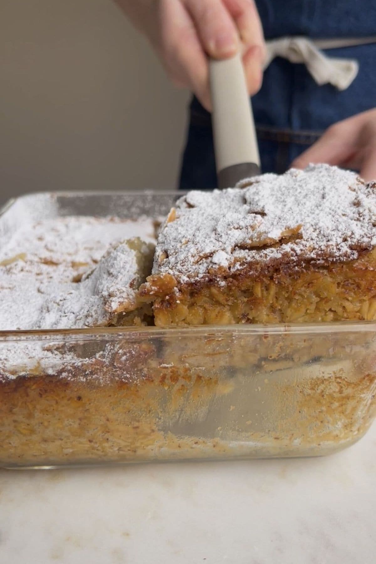 Spatula lifting a baked oat square dusted with powdered sugar from a glass baking dish.