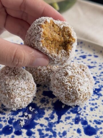 Hand holding a bitten coconut-coated ball above more on a blue speckled plate.